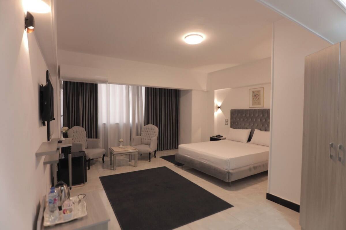 A modern hotel room with a bed, a desk, two chairs, a TV, and a large window with curtains.