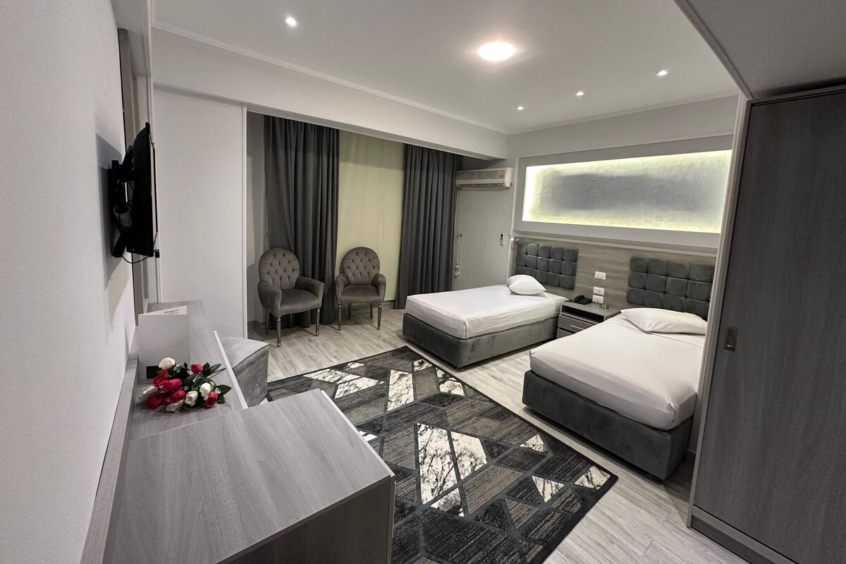 A modern hotel room with two beds, a desk, a TV, and a large window.