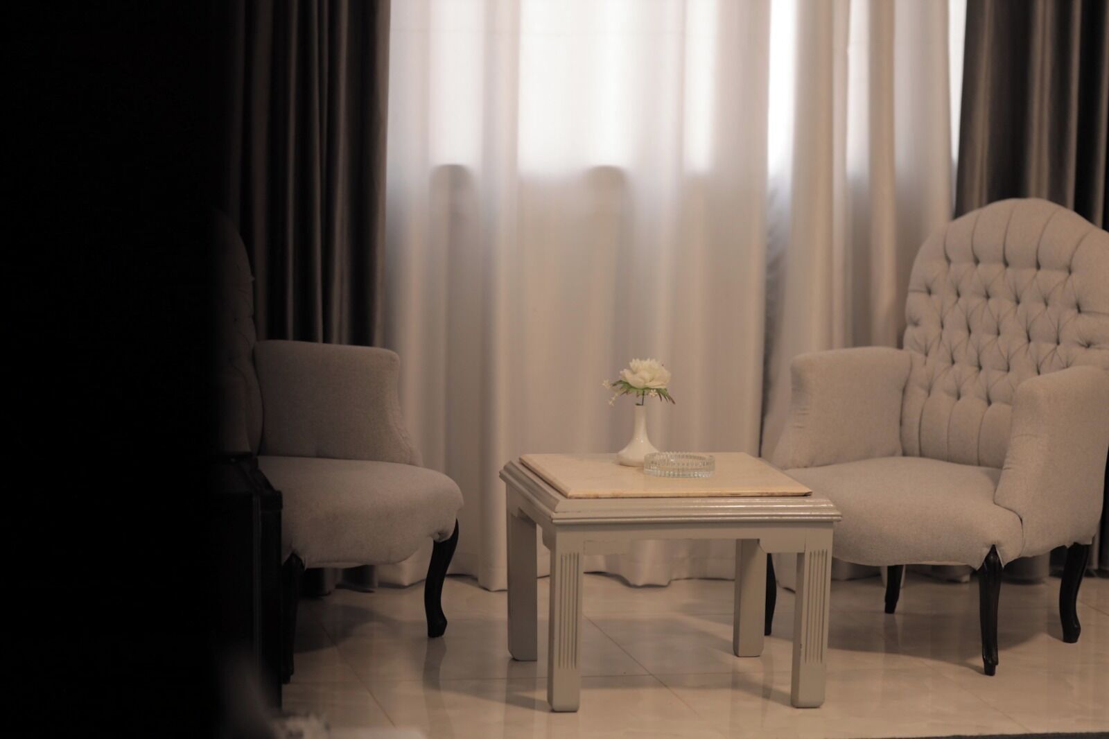 A room with two armchairs and a small table, a vase with flowers, and a view through sheer curtains.