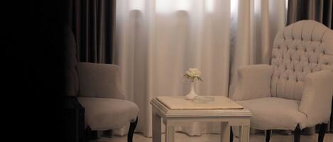 A room with two armchairs and a small table, a vase with flowers, and a view through sheer curtains.