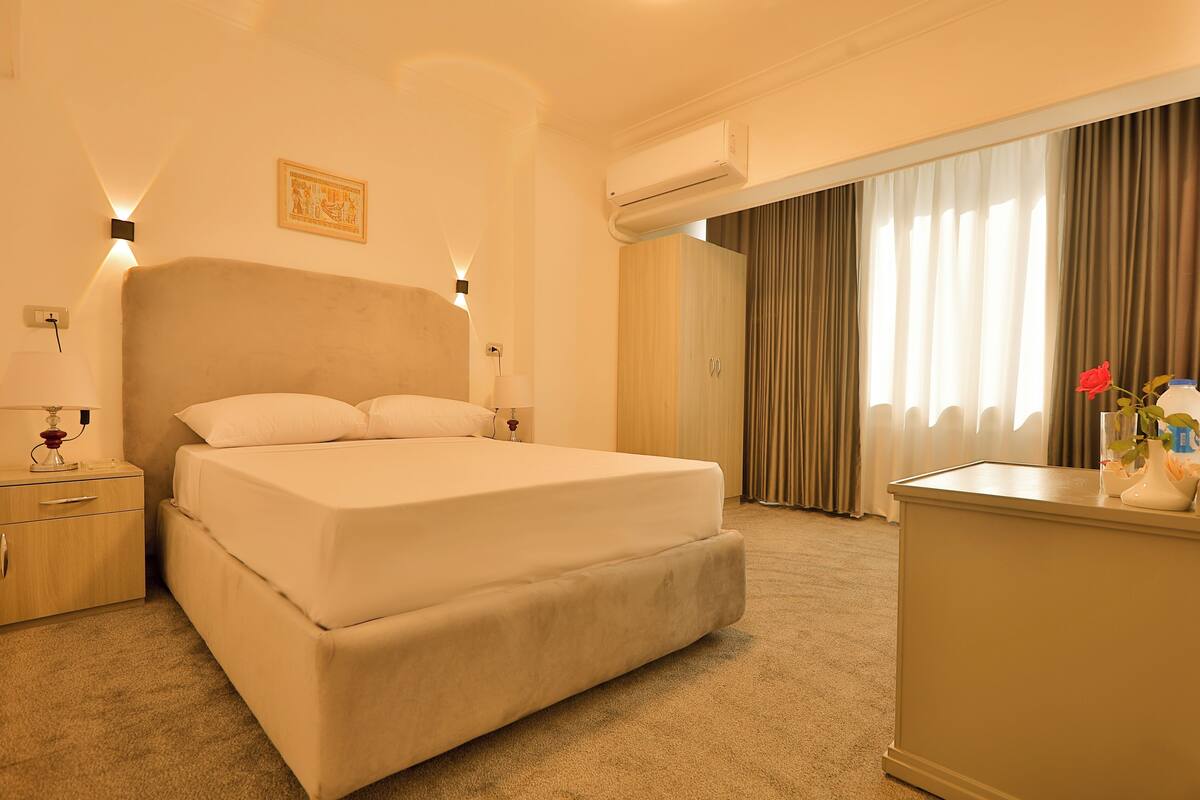 A hotel room with a large bed, bedside lamps, an air conditioning unit, a desk with a vase of flowers, and a window with curtains.