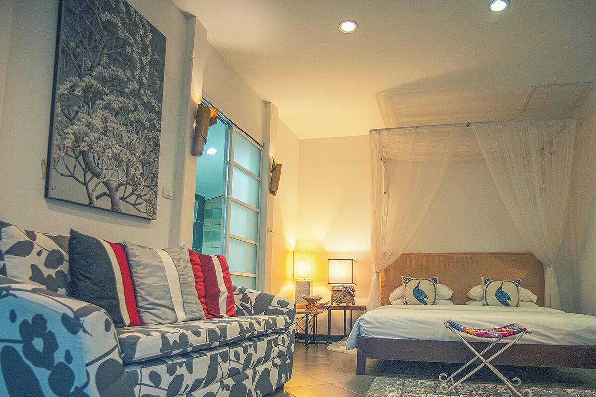 Un dormitorio con cama, sofá, mesita de noche, lámpara y ventana con cortinas.