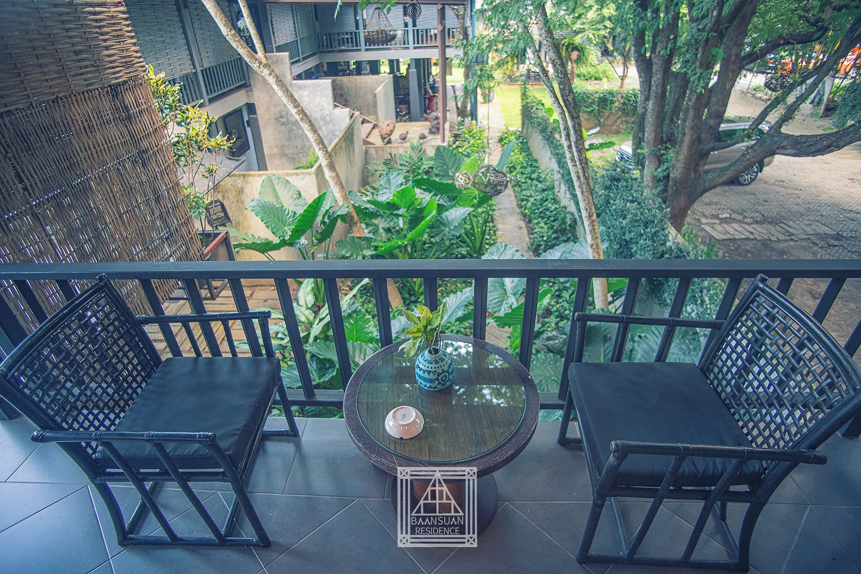 A balcony with two black wicker chairs, a small table with a vase, and a sign reading 'BAANSUAN RESIDENCE'.