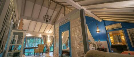 Una habitación amplia con techo alto, un ventanal con cortinas, una cama, un espacio de descanso con un sillón estampado, y una pared decorativa con detalles en azul.