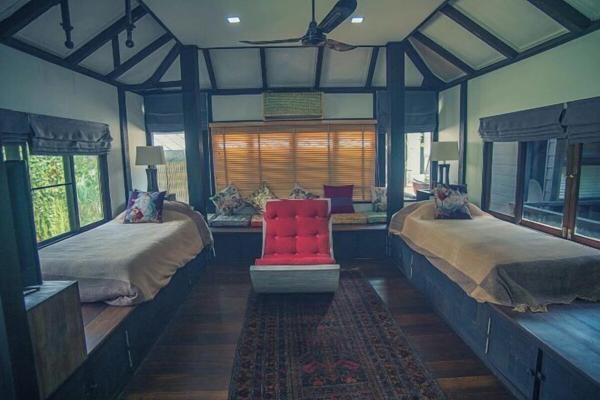 A room with a high ceiling, a central red chair, two beds with headboards, a large window with blinds, and a ceiling fan.