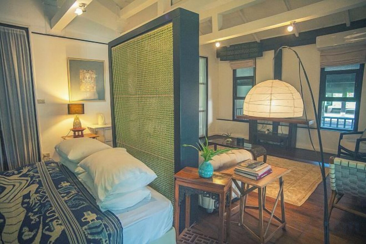 A room with a bed, a small table with a lamp, a chair, and a large window.