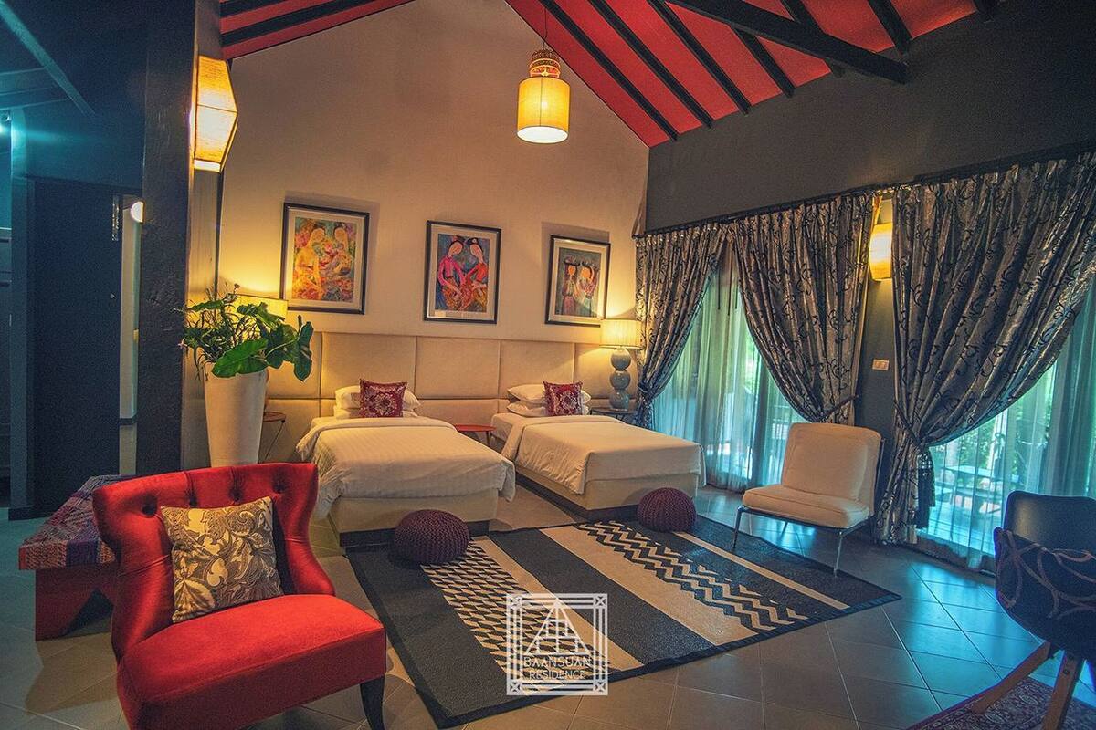 Habitación de hotel con dos camas, un sillón rojo, un sofá blanco, una planta grande en maceta y obras de arte en las paredes.