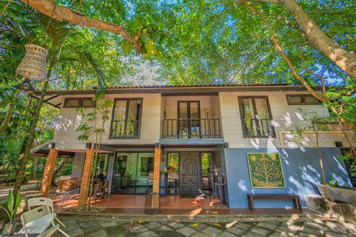 Un edificio de dos pisos con balcón, rodeado de árboles y un patio con mobiliario exterior.