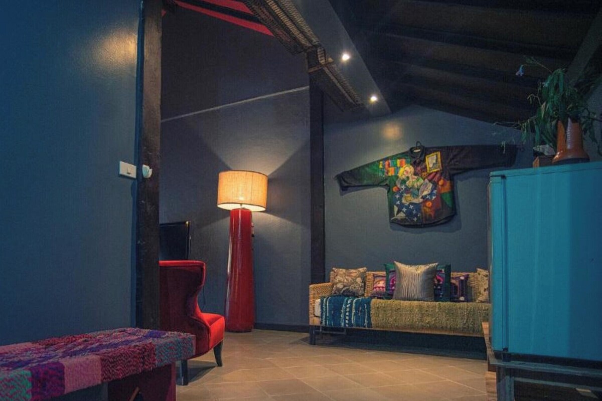A room with a red chair, a sofa, a table with a patterned tablecloth, a floor lamp, and a wall with a colorful artwork.