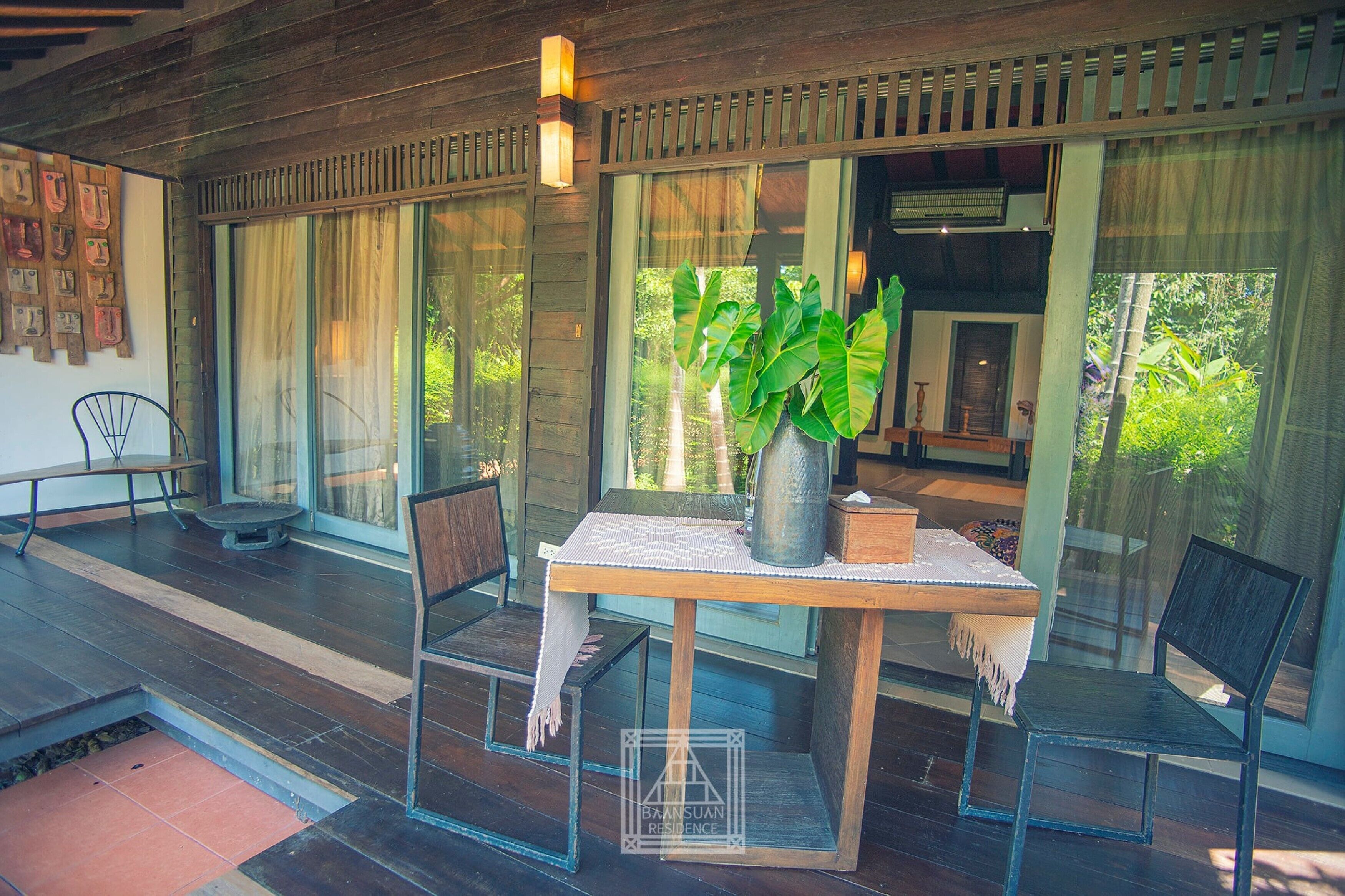 A wooden deck with a table and chairs, a potted plant, and a view of greenery through large glass doors.