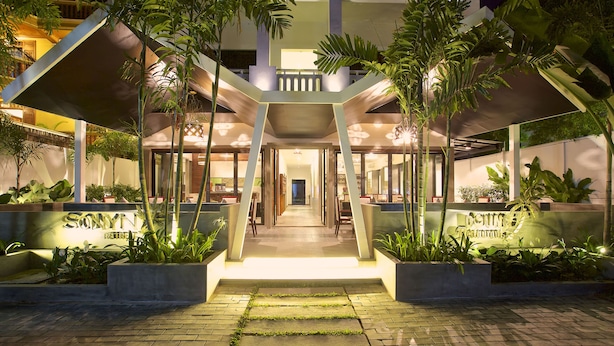 Entrada de hotel com uma escadaria central, cercada por floreiras e um caminho bem iluminado.