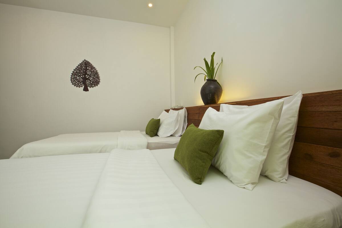 Ein Hotelzimmer mit zwei Betten, weißer Bettwäsche, grün-weißen Kissen, einem hölzernen Kopfteil, einer Wandpflanze und einem Wandbild.