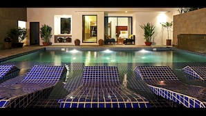 Piscina com espreguiçadeiras azulejadas e, através das portas de vidro, um quarto com cama e área de estar visível.