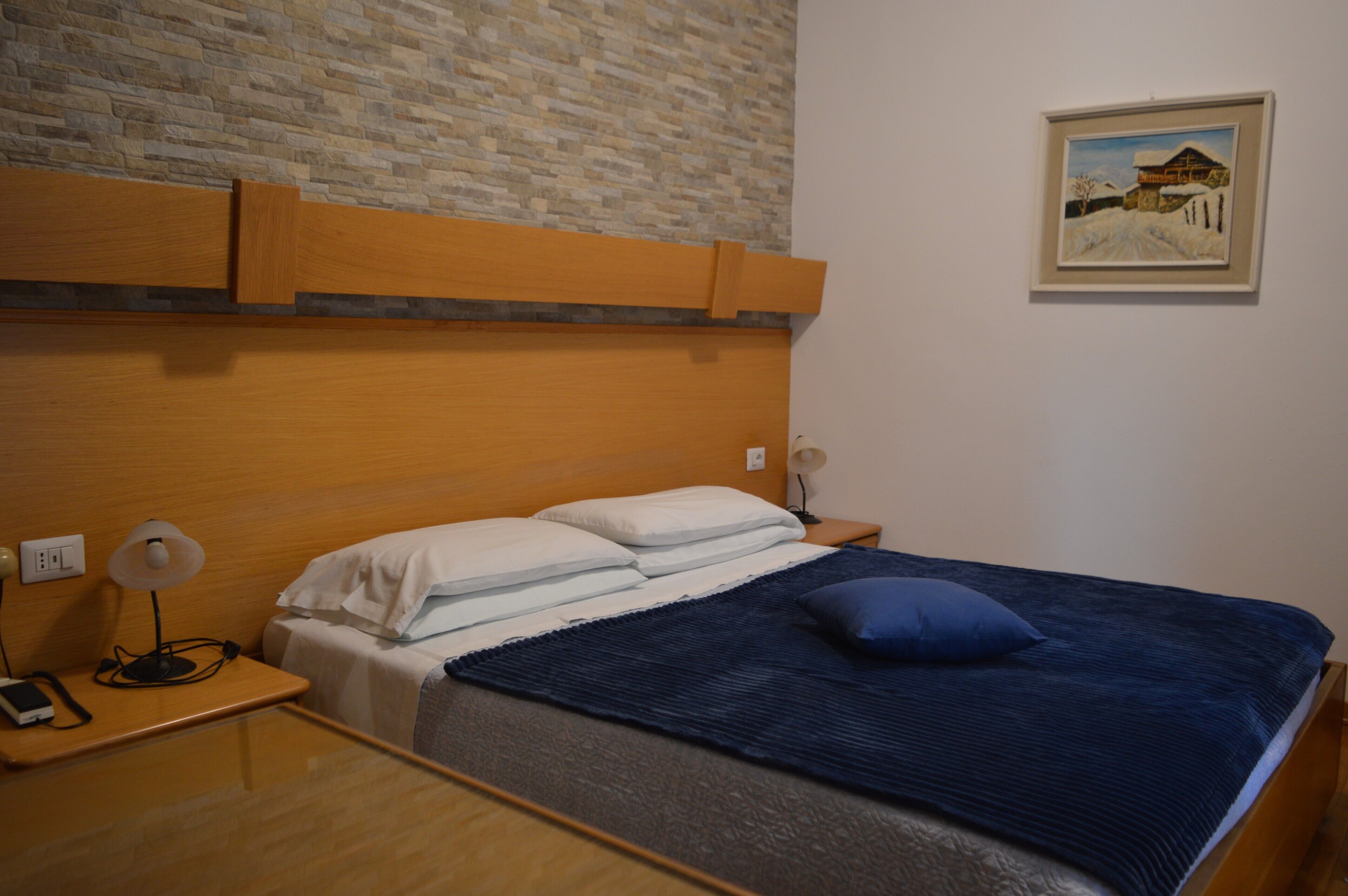 A hotel room with a wooden headboard, a bed with white and blue bedding, a nightstand with a lamp, and a painting on the wall.
