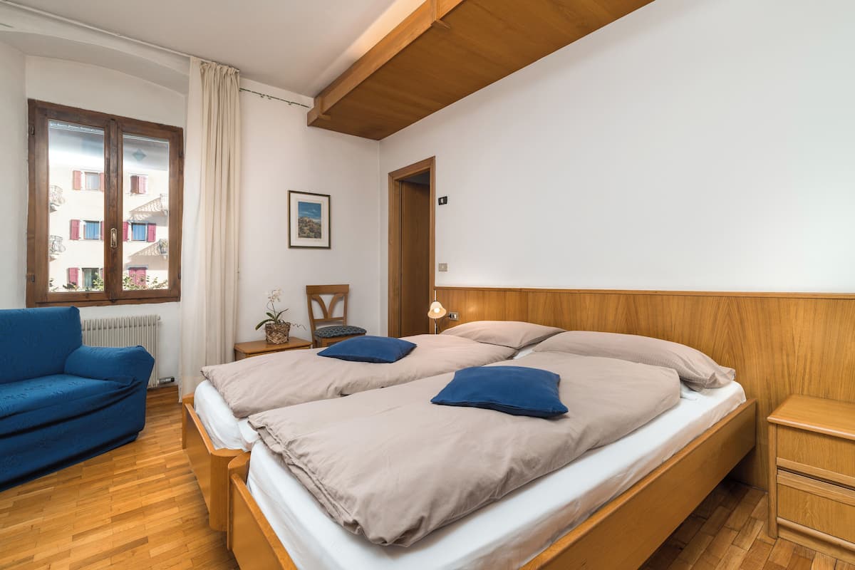 A bedroom with a large bed, a blue sofa, a wooden headboard, a nightstand, and a window with a view of buildings.