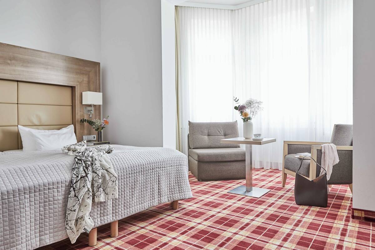 A hotel room with a bed, a chair, a small table, and a window with curtains.