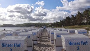 Filas de habitaciones de hotel blancas y numeradas, con la etiqueta 'Binzer Hof', frente a una playa, bajo un cielo despejado y colinas distantes.
