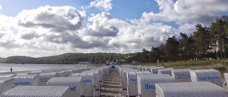 De nombreuses chambres d’hôtel blanches, numérotées et portant l’inscription « Binzer Hof », se dressent sur fond de plage, avec un ciel dégagé et des collines lointaines.