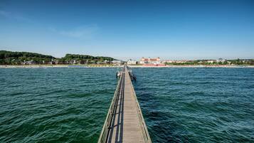 Une jetée en bois s’étendant sur une mer calme, menant à une ville côtière dont les bâtiments et la végétation se dessinent à l’arrière-plan.
