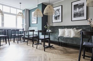 A modern dining area with a wooden herringbone floor, dark wooden chairs, and a green sofa with white cushions. There are two hanging woven light fixtures and framed black-and-white photographs on the wall. - Hotel Binzer Hof (Binz)