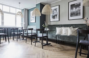 Ein moderner Essbereich mit Holzboden im Fischgrätmuster, dunklen Holzsesseln und einem grünen Sofa mit weißen Kissen. Zwei hängende gewebte Leuchten und schwarz-weiße Bilderrahmen schmücken die Wand.