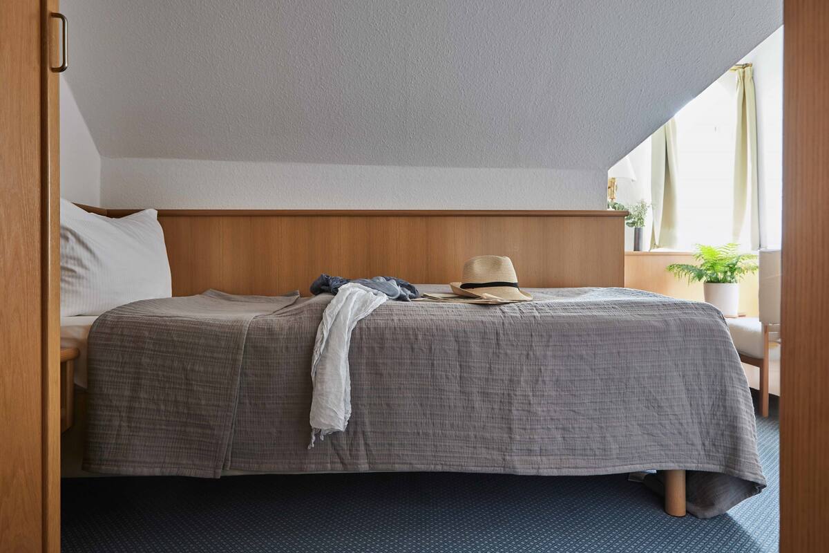 A neatly made bed with a grey blanket, a white pillow, and a hat on the headboard. There is a small table with a lamp and a plant beside the bed.