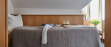 A neatly made bed with a grey blanket, a white pillow, and a hat on the headboard. There is a small table with a lamp and a plant beside the bed.