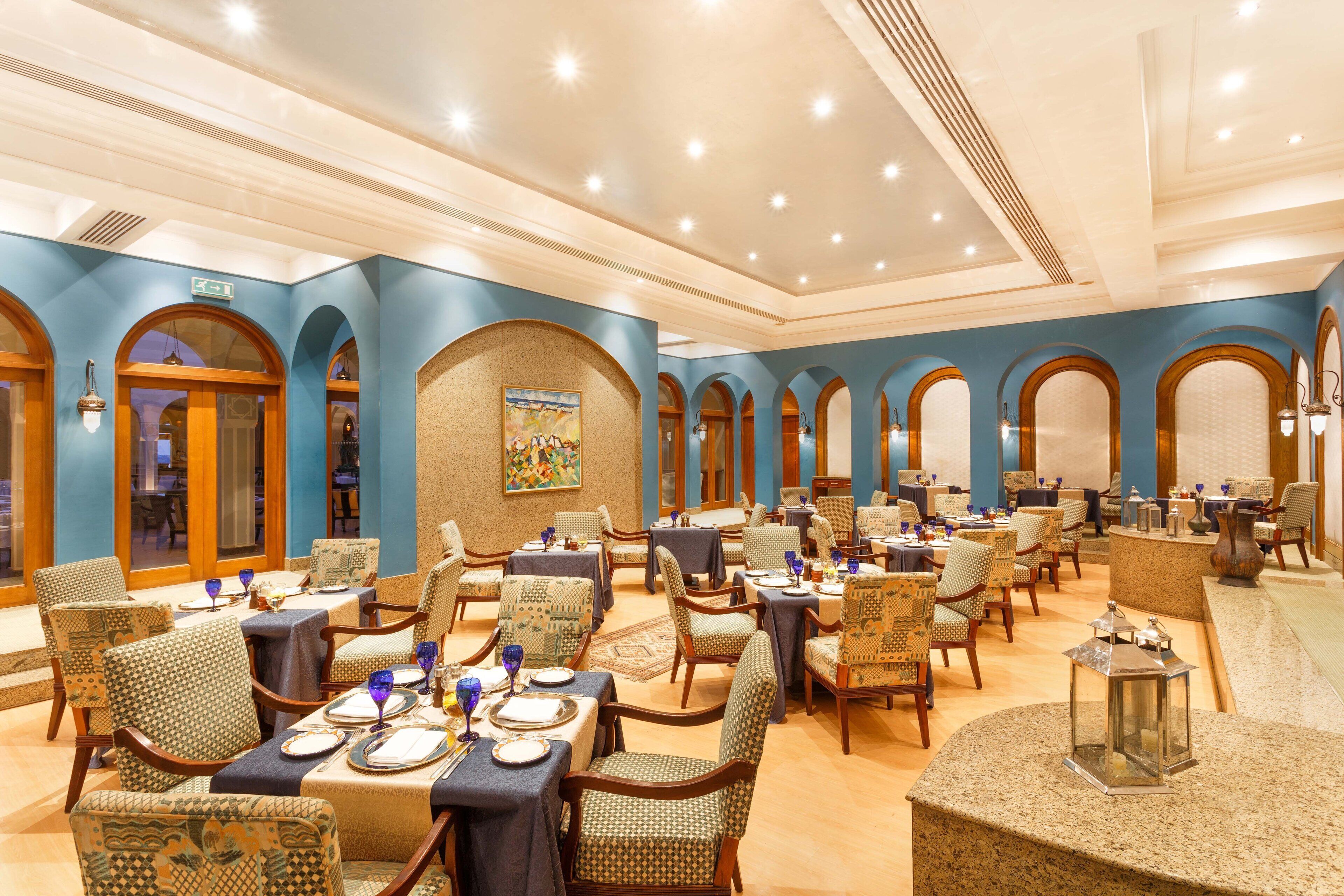 A spacious dining area with multiple tables and chairs, featuring a classic design with arched doorways and a central mural.