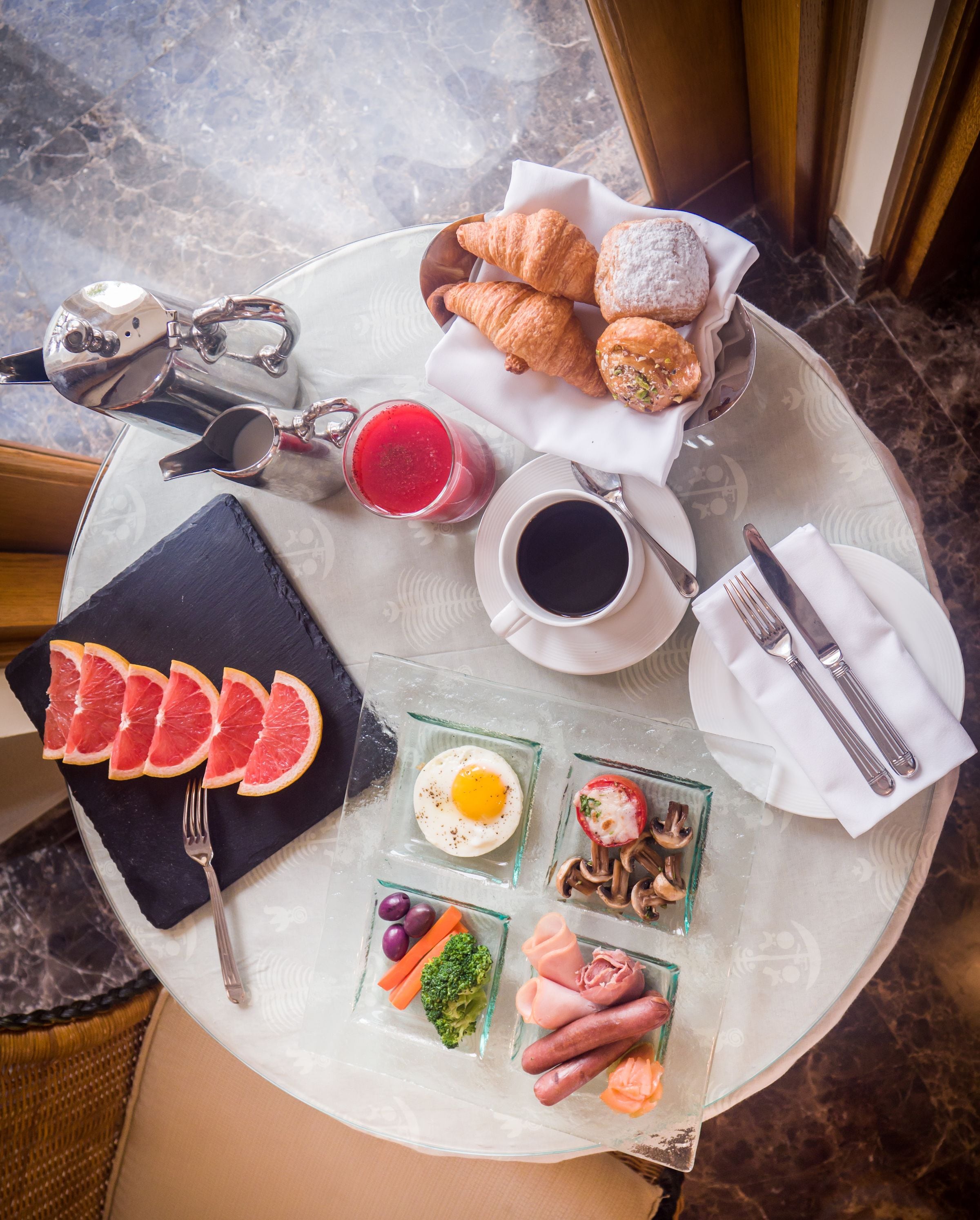 A breakfast setting with croissants, a glass of red juice, a cup of coffee, and various breakfast items on a glass table.