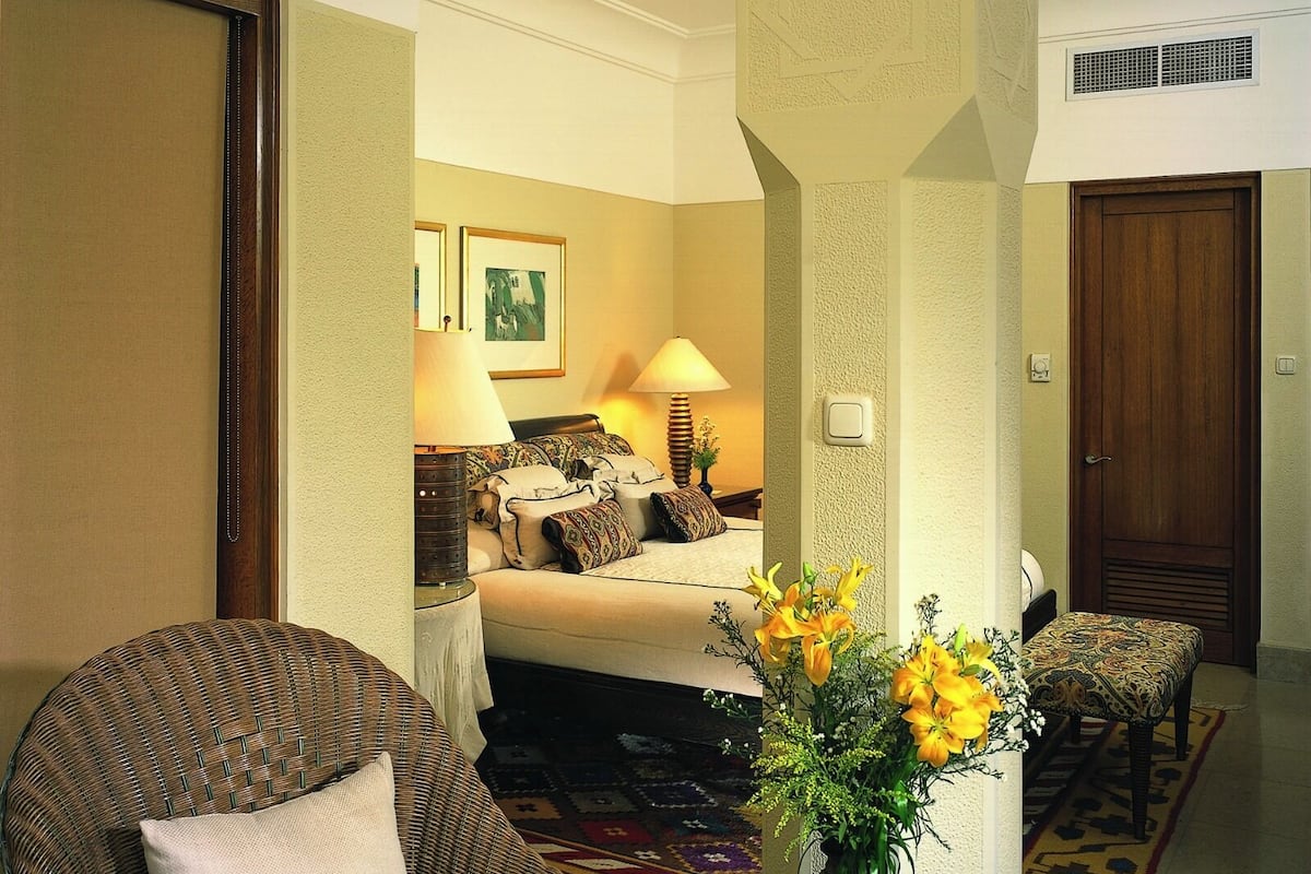 A hotel room with a bed, a sitting area with a chair and a table, a vase with flowers, and a door.