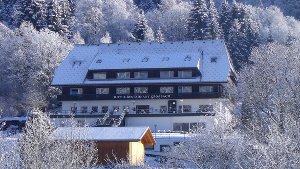 Un hôtel dont le toit est recouvert de neige, qui possède de nombreuses fenêtres et une enseigne bien visible indiquant « HÔTEL RESTAURANT GROOCH ».