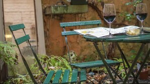 A green metal table with two wine glasses and a bowl, set against a brick wall with climbing plants and a hanging flower pot.