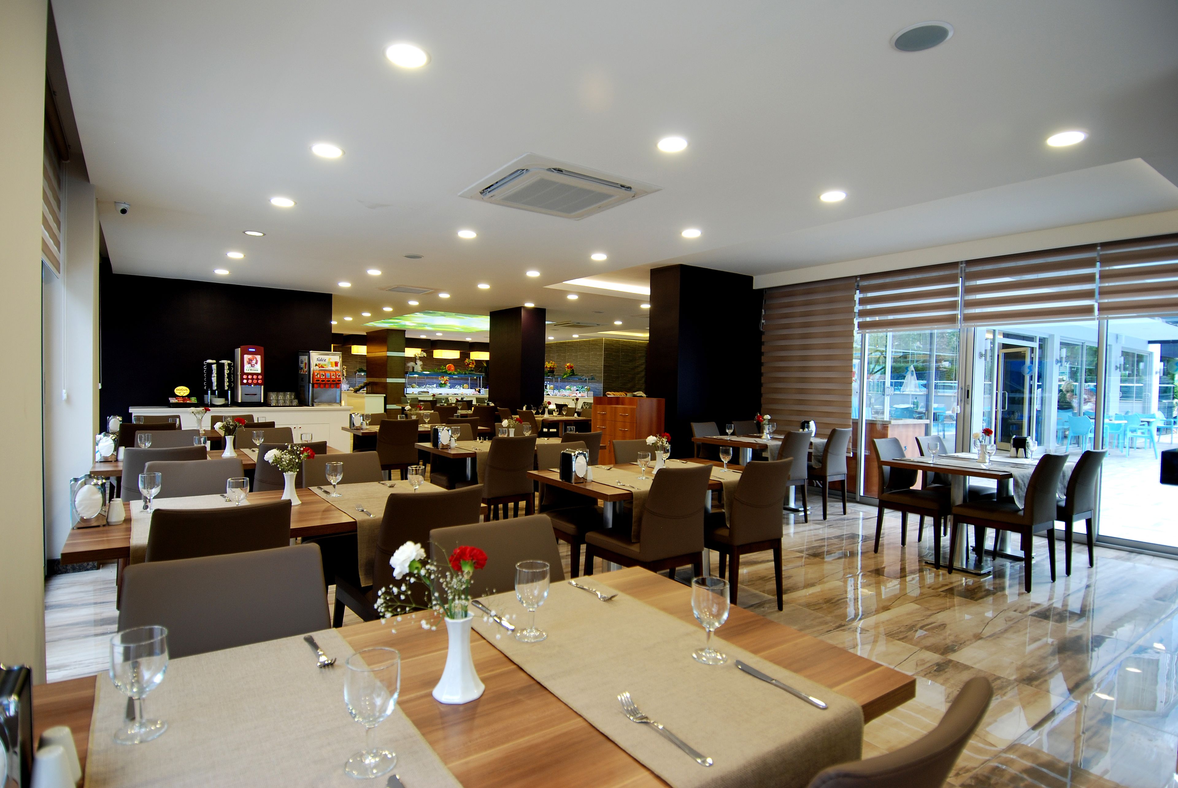 A spacious restaurant with multiple dining areas, a visible kitchen, and large windows offering a view of the outdoors.