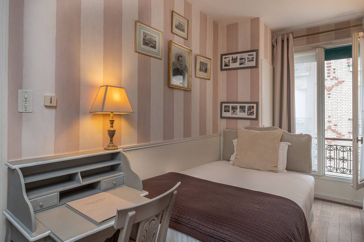 A hotel room with a bed, desk, chair, lamp, and a balcony with a view of buildings.
