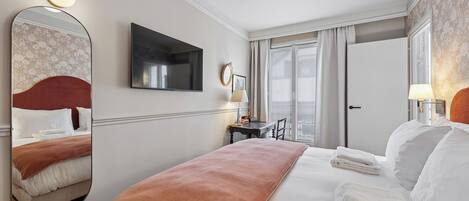 A hotel room with a bed, a flat-screen TV, a large mirror, a desk with a lamp, and a view of the outdoors through a window.