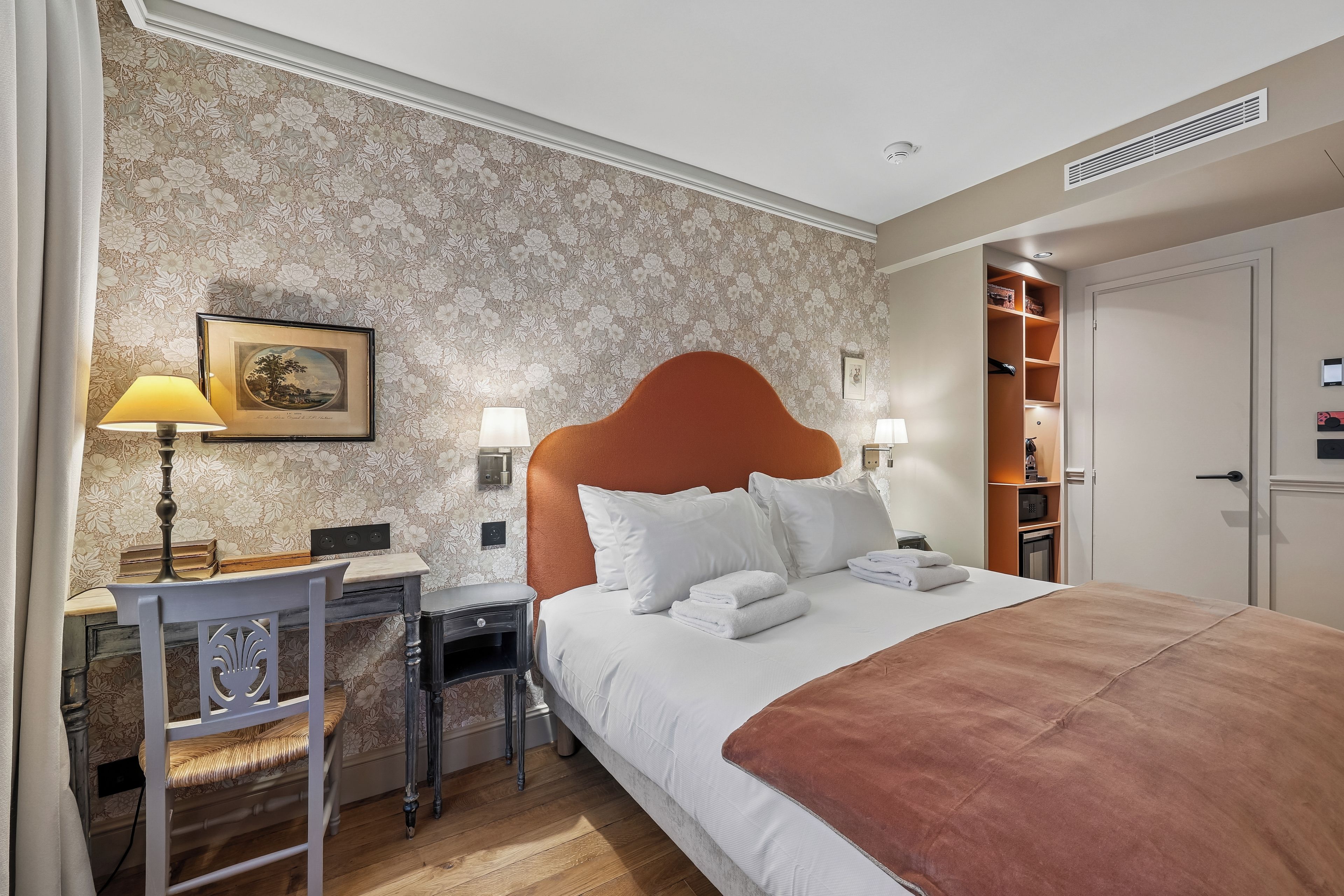 A hotel room with a bed, bedside table, lamp, and a wall with floral wallpaper.