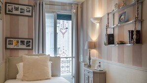A hotel room with a bed, a nightstand, a lamp, a shelf with books and decorative items, and a view of a brick wall with a heart-shaped window. - Hotel de l'Avre (Paris)