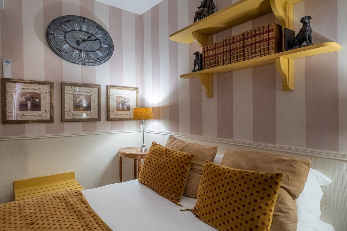 A hotel room with a bed, pillows, a clock, framed pictures, a lamp, and a shelf with books and a statue.