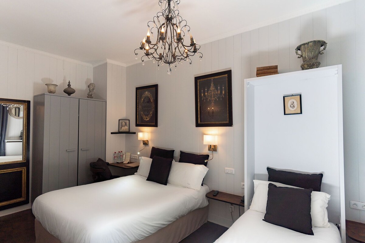 A hotel room with a large bed, bedside tables, a wardrobe, a chandelier, and wall-mounted lamps.