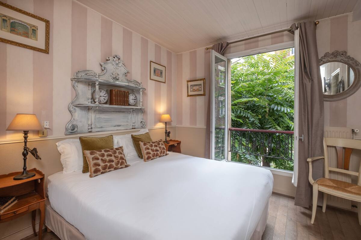 A hotel room with a large bed, two bedside lamps, a wooden nightstand, a chair, a mirror, and a balcony with greenery.