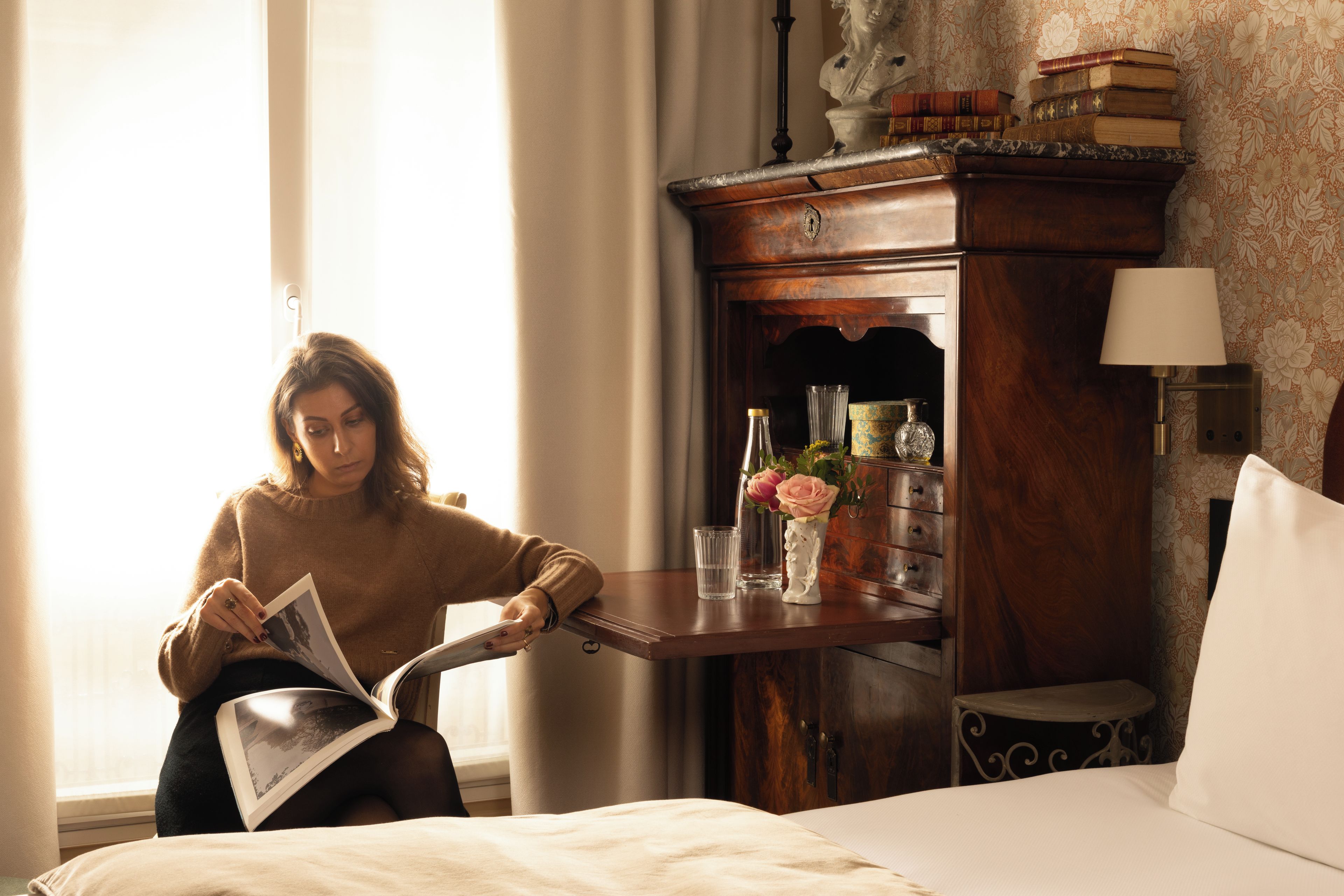 Une personne est assise dans une pièce, lisant un livre. On y voit un lit, une armoire en bois remplie de livres et un vase avec des fleurs.