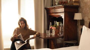 Une personne est assise dans une pièce, lisant un livre. On y voit un lit, une armoire en bois remplie de livres et un vase avec des fleurs.