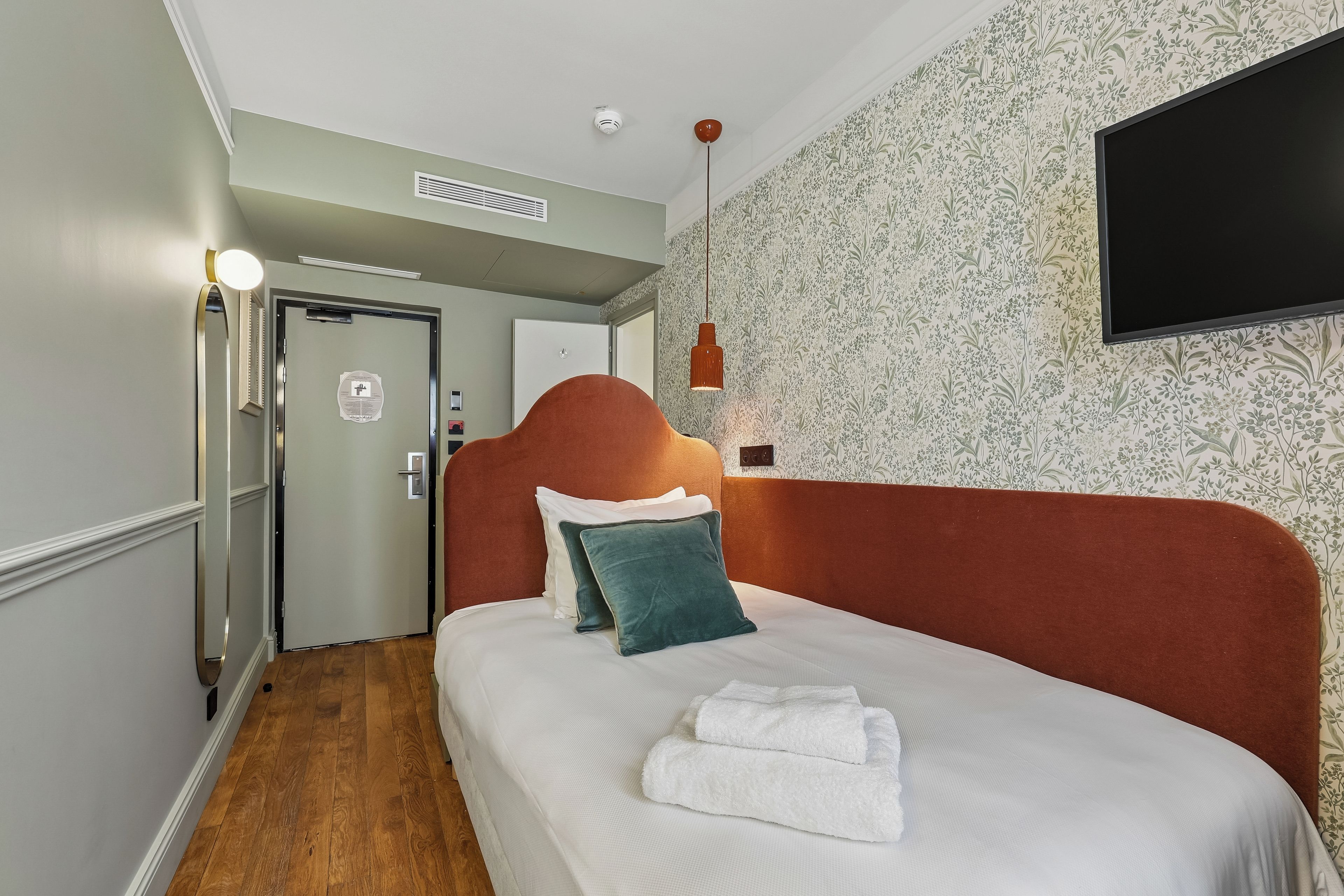 Ein Hotelzimmer mit Bett, einem flachbildfernseher an der Wand, einer Tür mit Überwachungskamera und Holzboden.