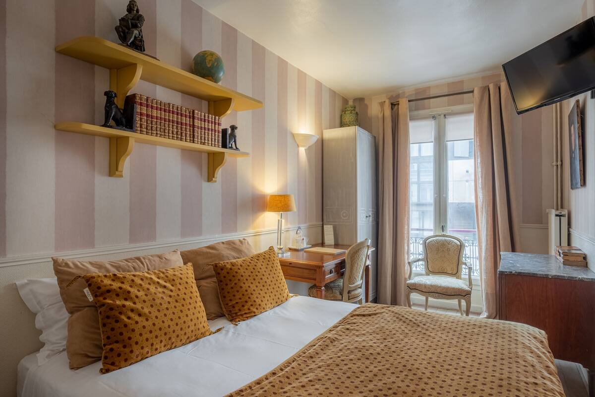 A hotel room with a bed, a desk, a chair, a TV, and a balcony with curtains.