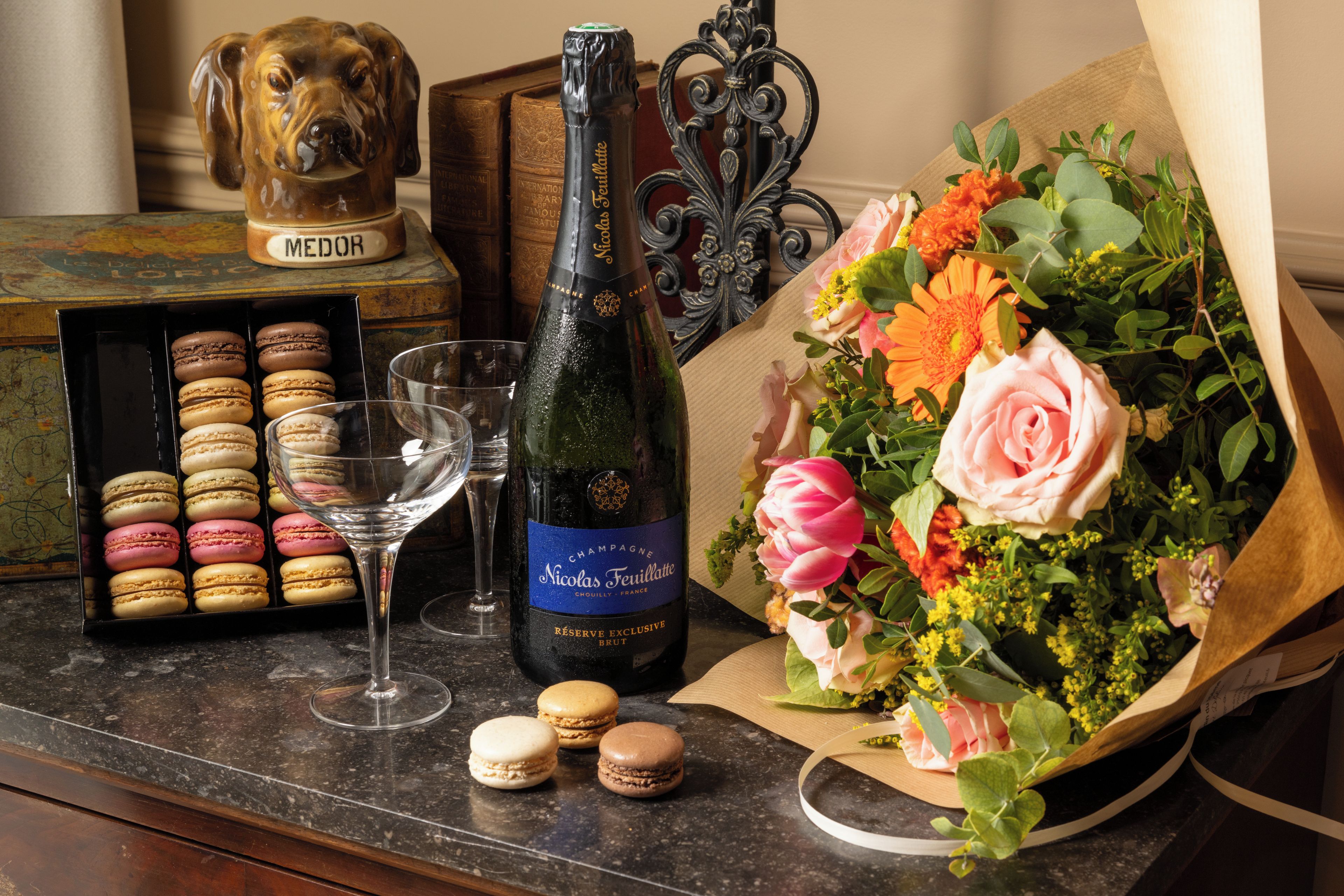 Une bouteille de champagne Nicolas Feuillatte, un verre de vin, des macarons et un bouquet de fleurs sur une table.