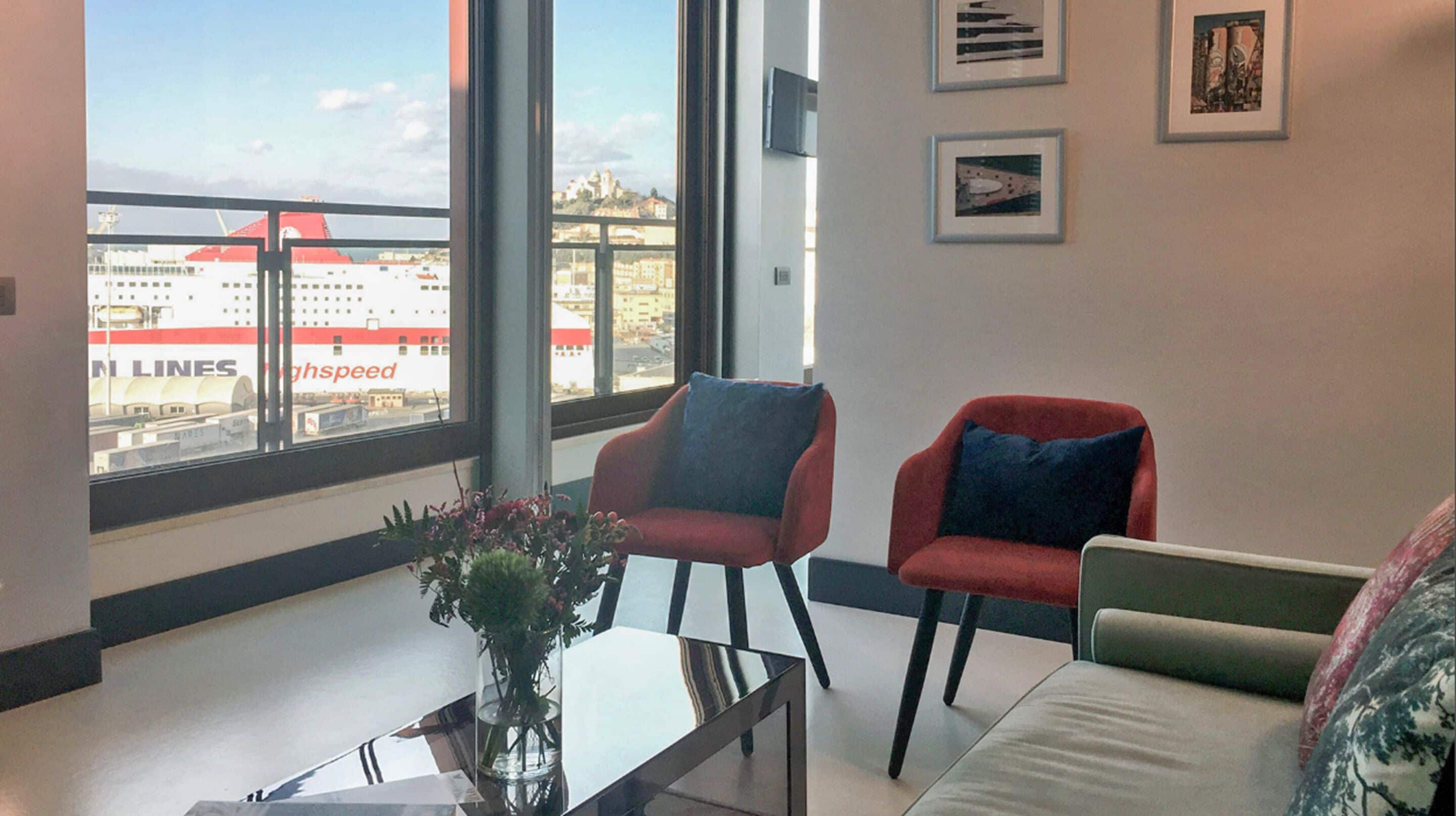 Una sala de estar moderna con un ventanal grande, dos sillones, un sofá y una mesa de centro con un jarrón de flores.