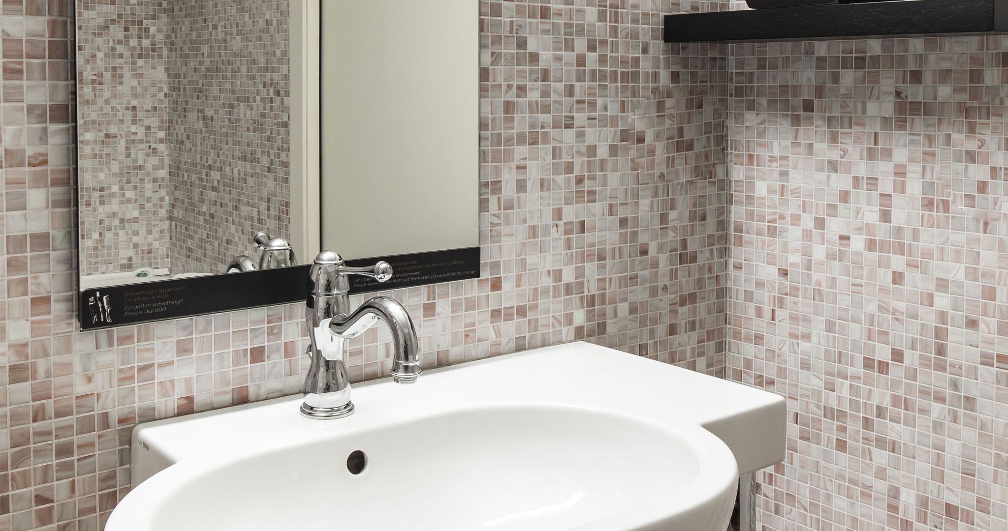 A bathroom with a white sink, a chrome faucet, and a mirror above it. The walls are tiled with a marble pattern.