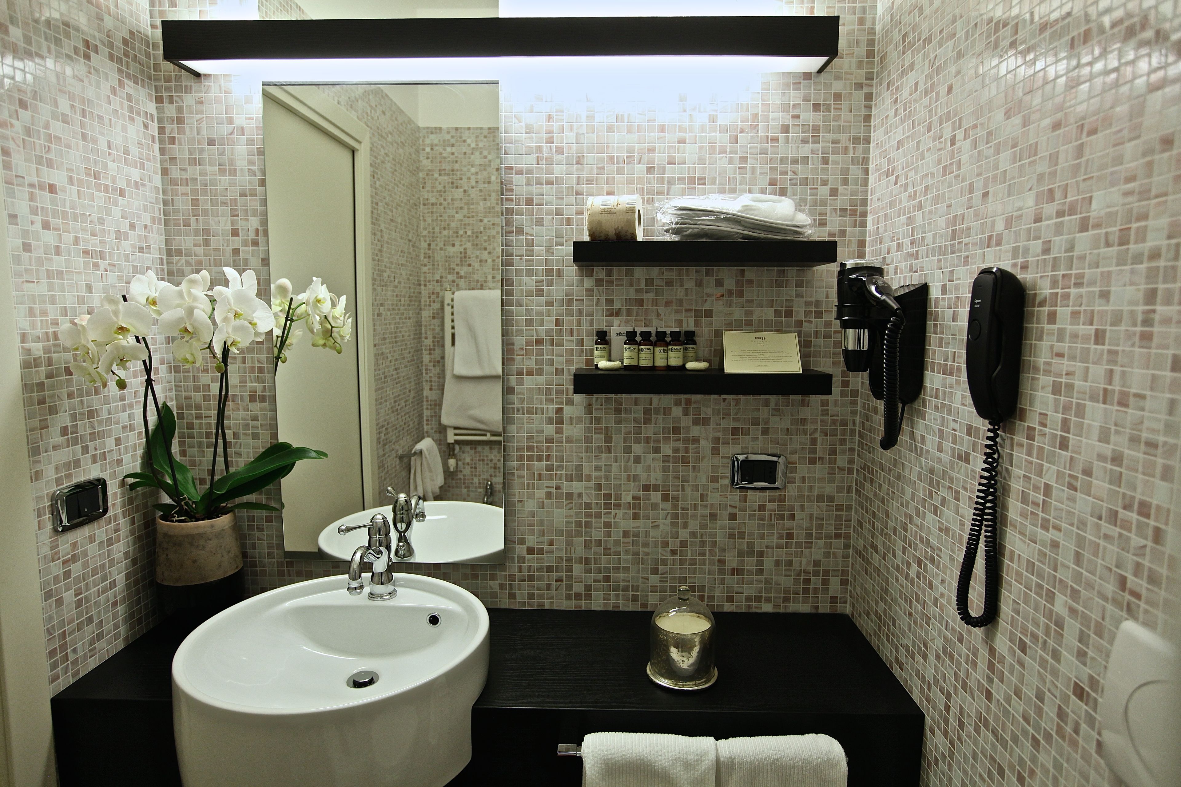 A bathroom with a white sink, a mirror, a potted orchid, a black countertop, a towel, a phone, and toiletries on a shelf.