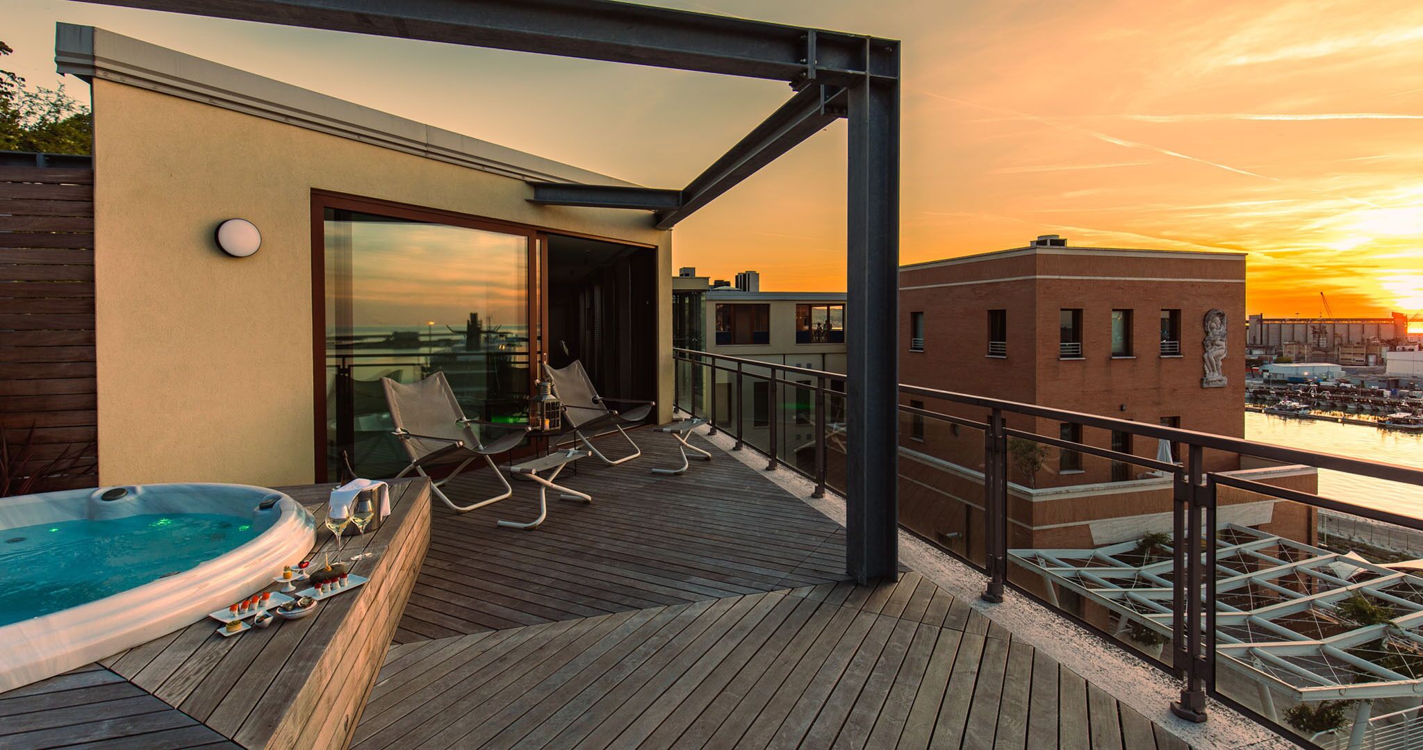 A rooftop terrace with a hot tub, lounge chairs, and a view of the sunset over a cityscape.