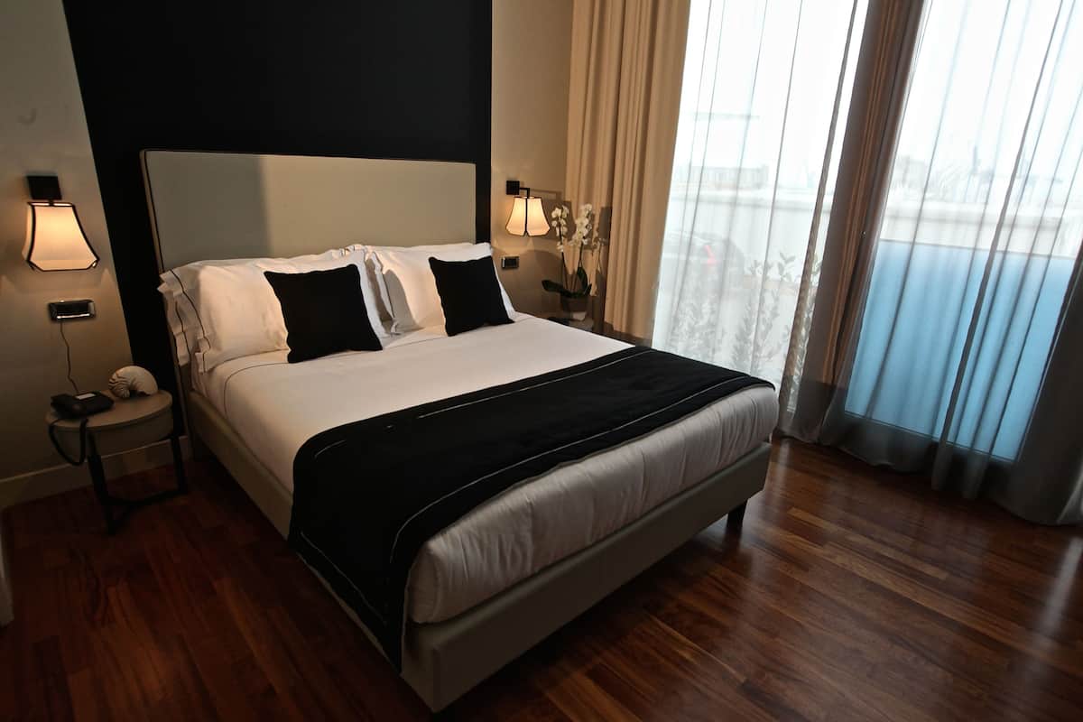 A hotel room with a large bed, a bedside table with a lamp, and a view of the outdoors through the window.